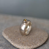 A handcrafted sterling silver ring with a wide, domed band and a polished finish. Set into the front of the band is a smooth, round light brown tourmaline gemstone in a bezel setting. The ring is photographed resting on a rough stone surface, highlighting its organic texture and contrast between the refined silver and earthy tones. The lighting emphasizes the gentle sheen of the silver and the warm depth of the gemstone.
