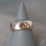 The image shows a handcrafted sterling silver ring with a wide, polished dome band. At its center is a smooth, round-cut light brown tourmaline gemstone, bezel-set for a clean, modern look. The silver surface is reflective and sleek, enhancing the warm, earthy tone of the tourmaline. The ring rests on a neutral background that highlights its shine and craftsmanship. The overall style is minimalist, elegant, and bold, ideal for women who love natural gemstone statement rings.