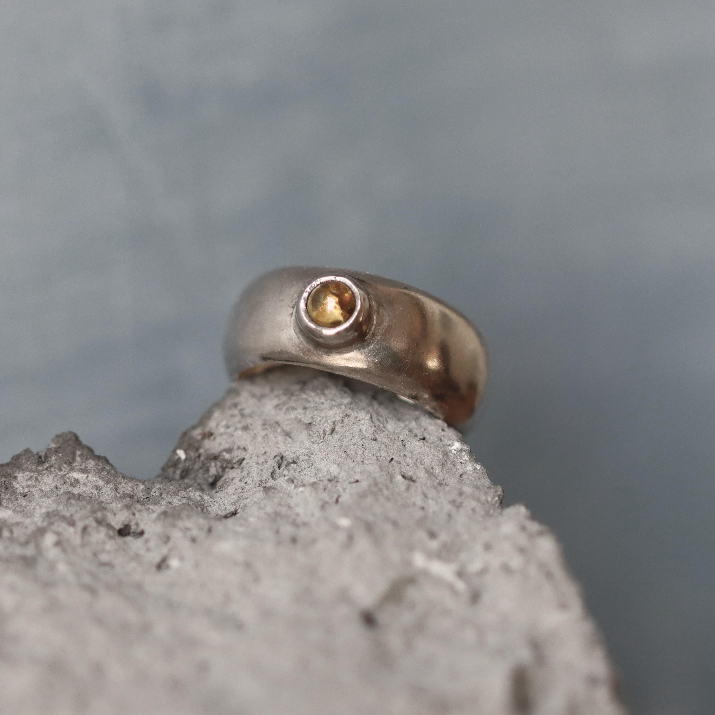 A handcrafted sterling silver ring with a wide, domed band and a polished finish. Set into the front of the band is a smooth, round light brown tourmaline gemstone in a bezel setting. The ring is photographed resting on a rough stone surface, highlighting its organic texture and contrast between the refined silver and earthy tones. The lighting emphasizes the gentle sheen of the silver and the warm depth of the gemstone.
