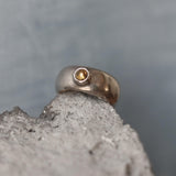 A handcrafted sterling silver ring with a wide, domed band and a polished finish. Set into the front of the band is a smooth, round light brown tourmaline gemstone in a bezel setting. The ring is photographed resting on a rough stone surface, highlighting its organic texture and contrast between the refined silver and earthy tones. The lighting emphasizes the gentle sheen of the silver and the warm depth of the gemstone.