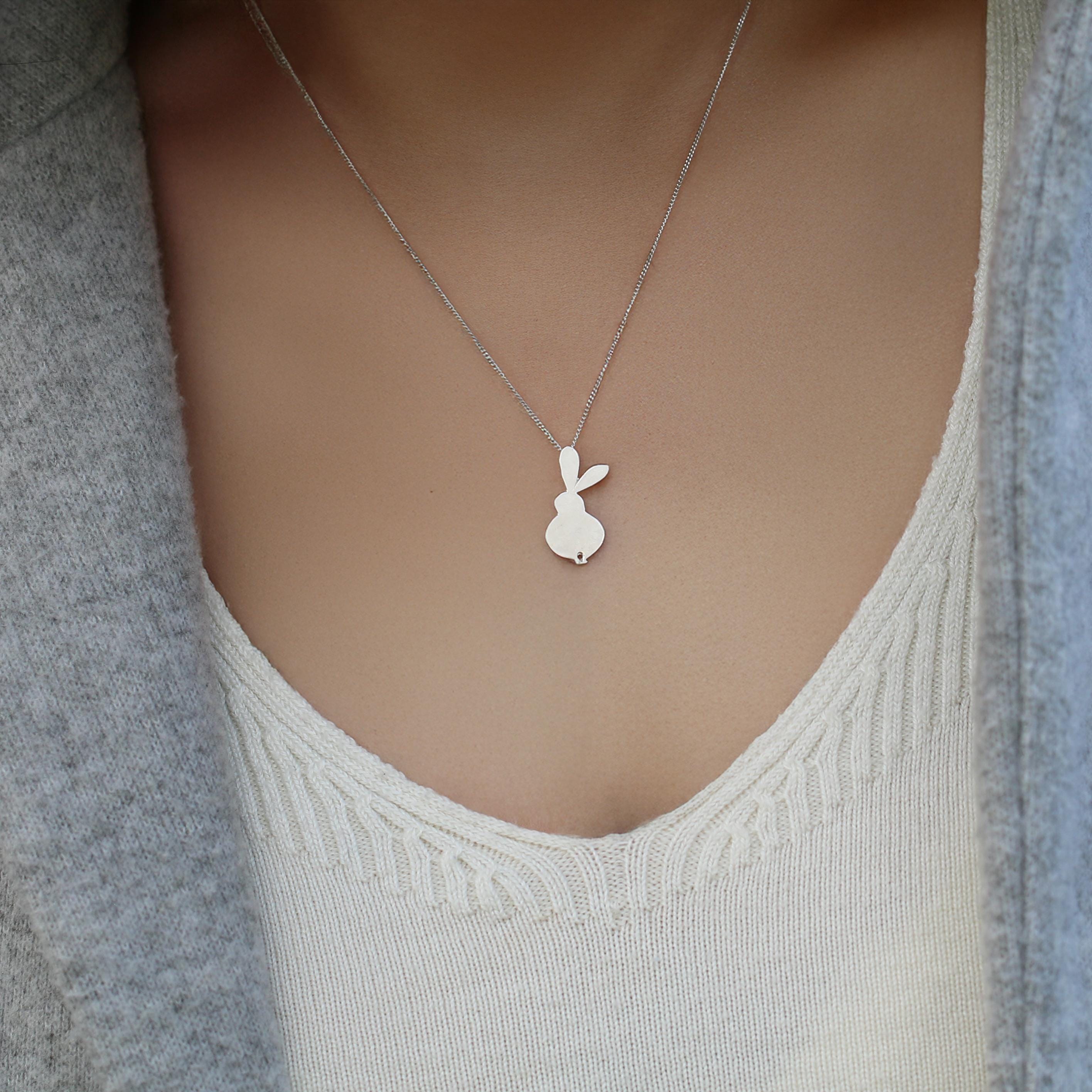 Close-up of a woman wearing a delicate sterling-silver necklace featuring a small minimalist bunny outline pendant centered on her chest, styled over a cream knit camisole and light gray cardigan.