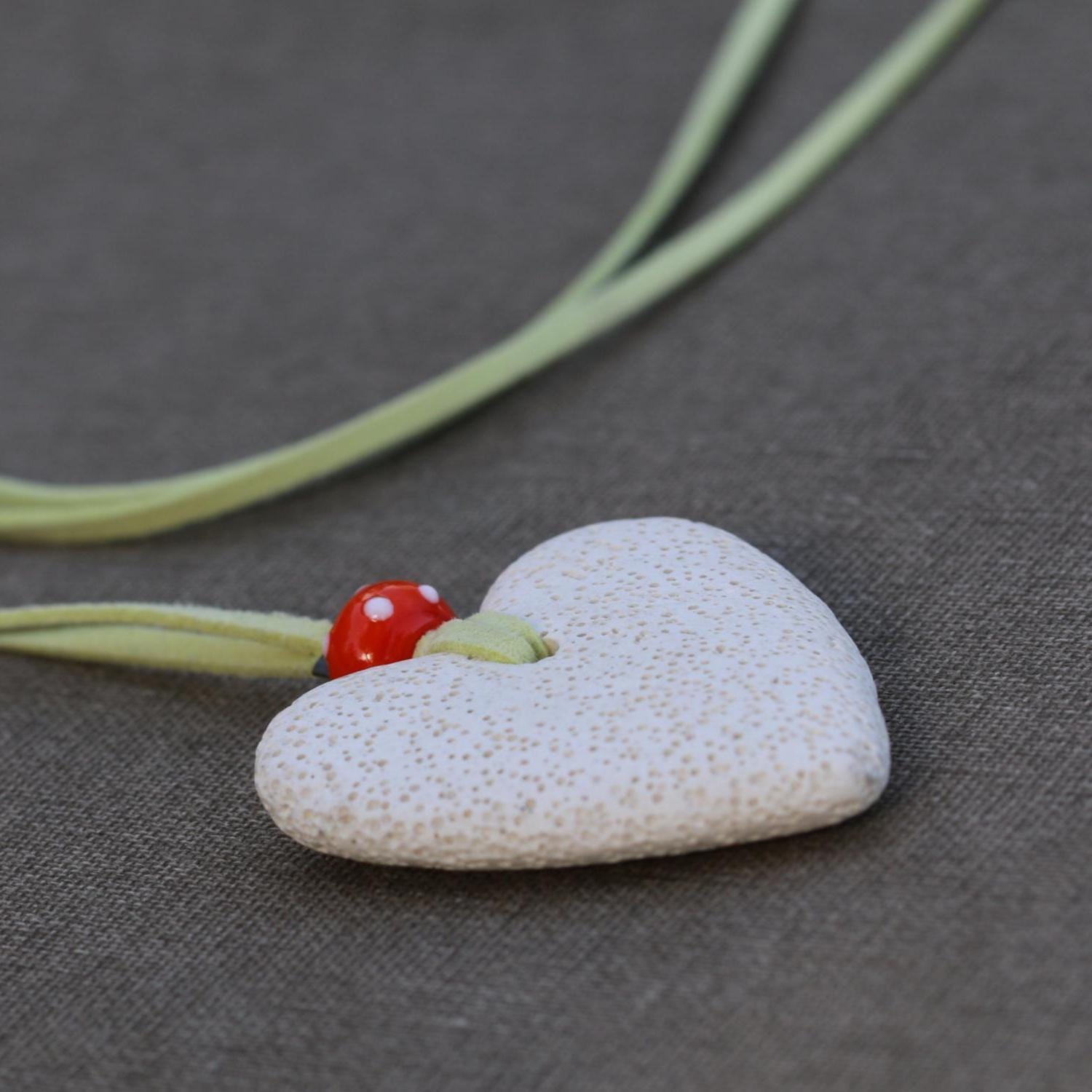 Artisan jewelry piece with a textured white lava heart pendant and orange glass charm, hanging from a long green suede cord.