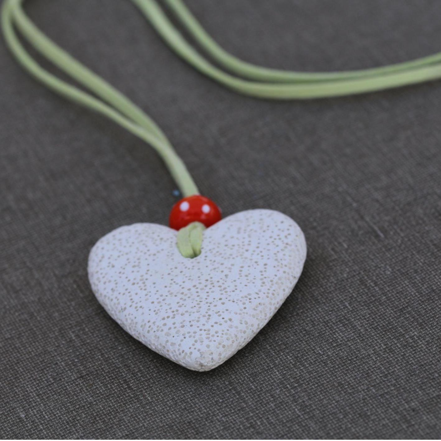 Detail shot of a white lava stone heart pendant necklace with a suede green strap and orange glass accent, photographed in natural light.