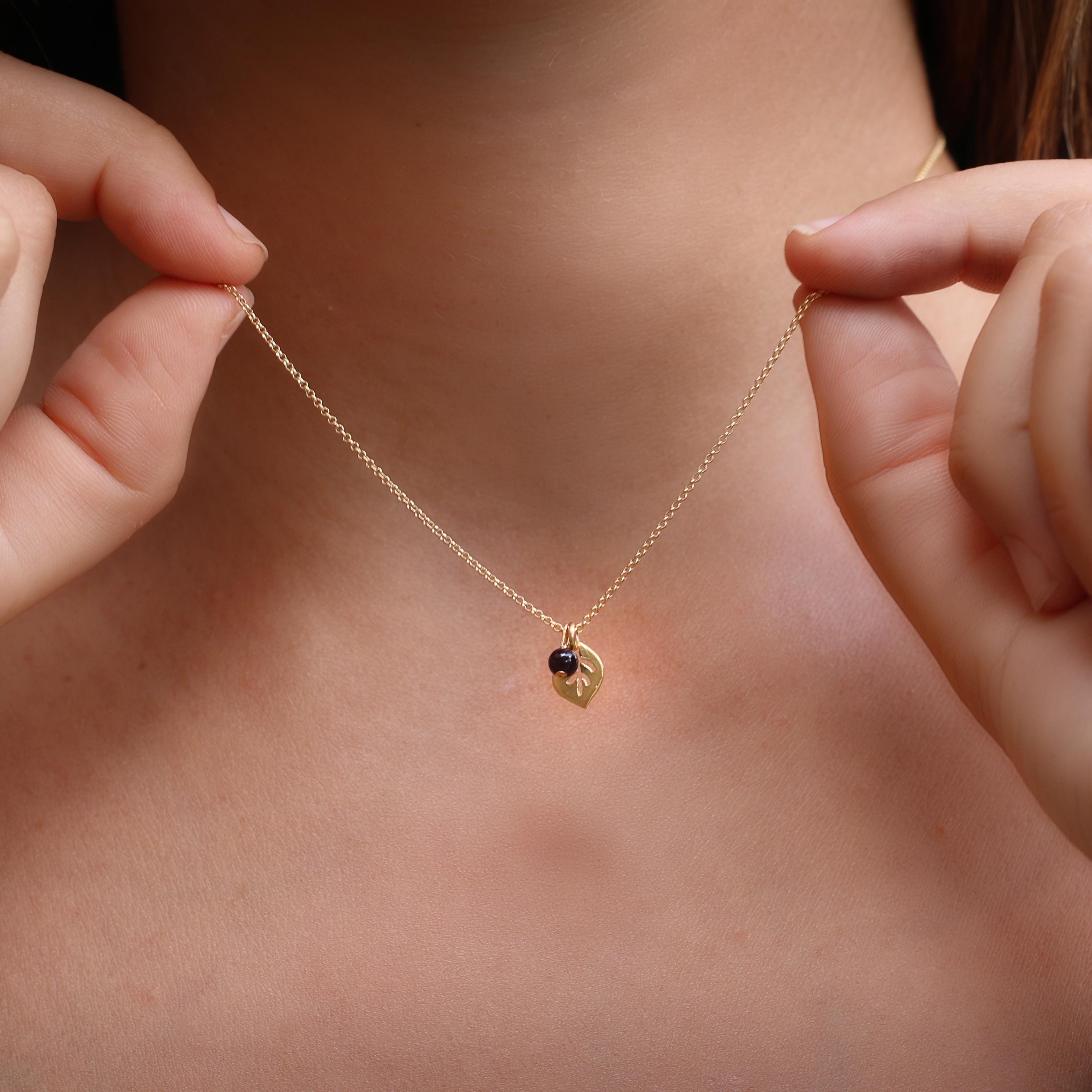 Model wearing a dainty gold leaf pendant with a petite garnet charm at the collarbone. The fine chain and small leaf silhouette create a subtle, feminine accent suitable for everyday wear or layering.