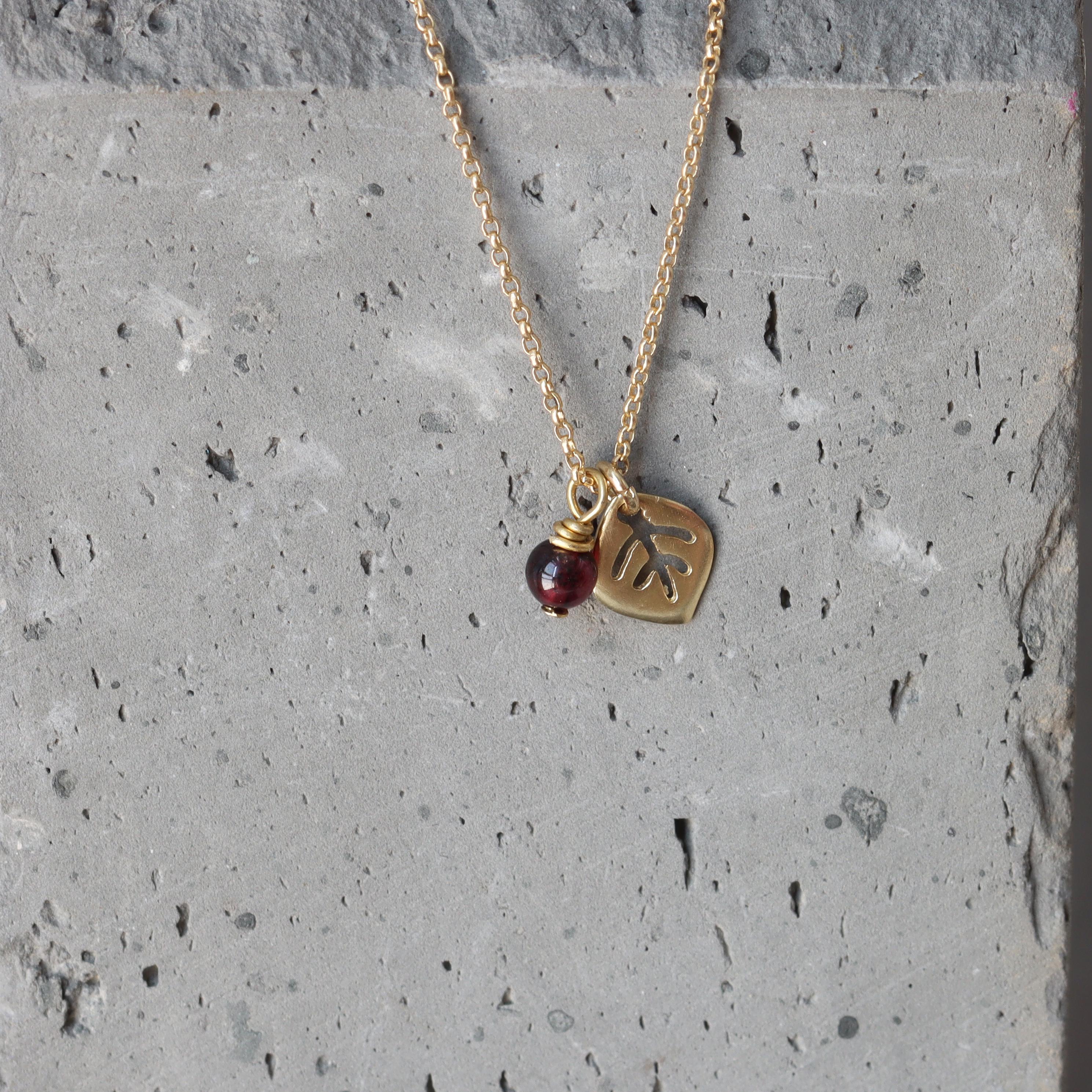 Macro image highlighting the pendantโs texture: fine leaf veins catch the light while a faceted garnet glows in deep crimson. The slim gold chain enhances the necklaceโs delicate, nature-inspired design.