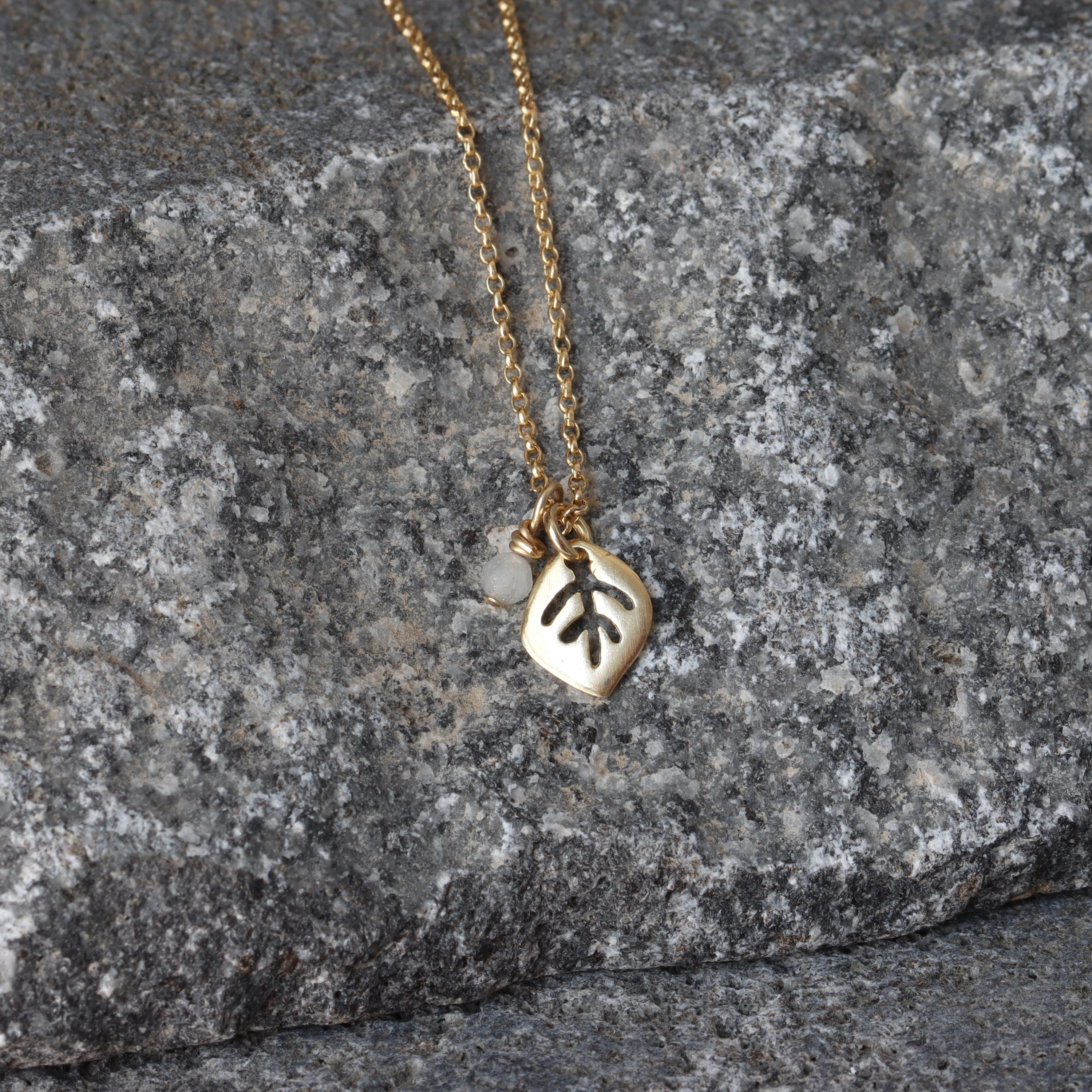 The necklace is arranged in a loose circle atop a matte gray stone surface. A small moonstone charm sits beside a satin-finished gold leaf pendant; the fine cable chain catches light subtly. The cool gray background makes the warm gold and opalescent gem stand out.
