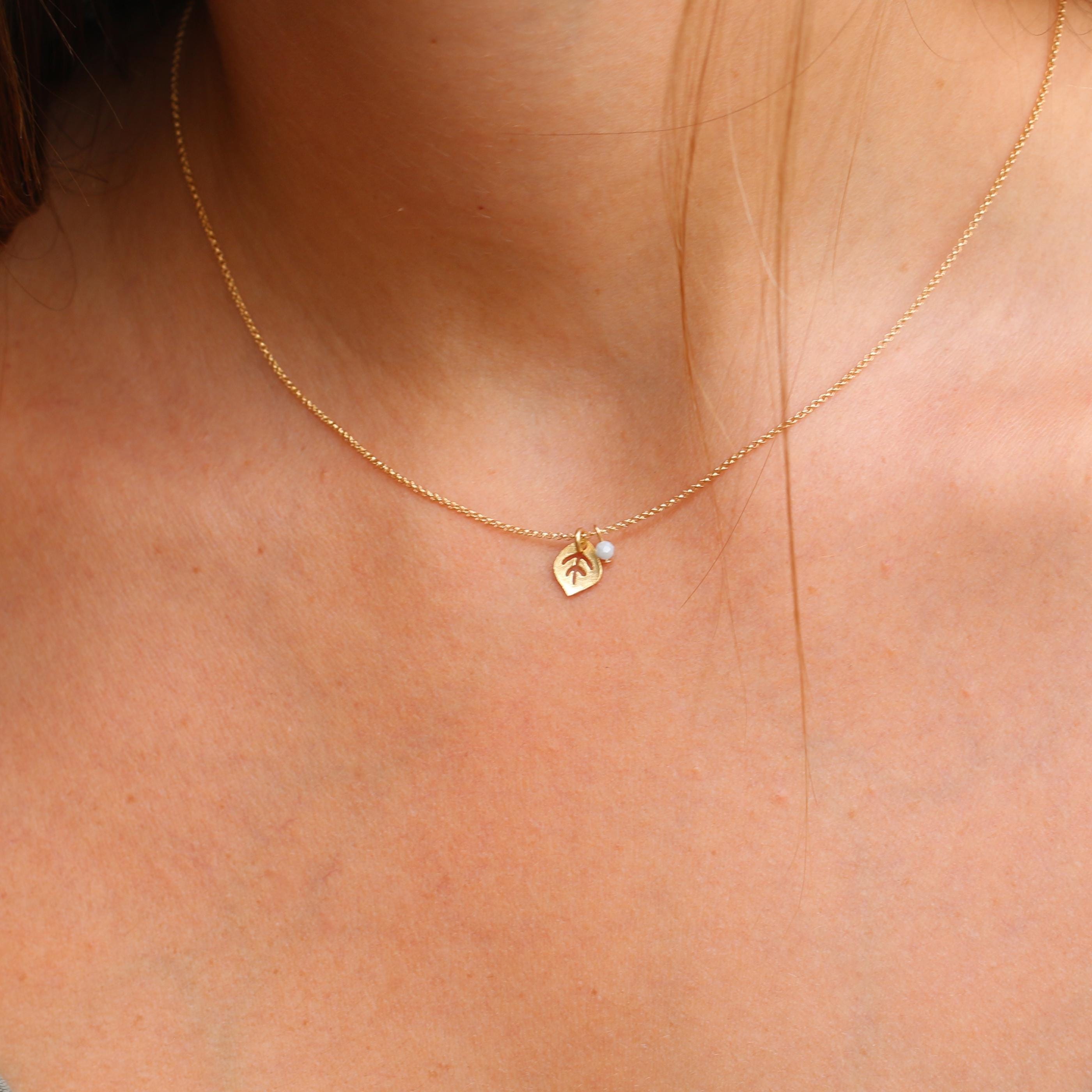 The gold leaf and moonstone birthstone necklace is styled over a simple blouse. The lightweight chain drapes naturally, with the leaf pointing down and the moonstone charm resting beside it, creating a soft botanical accent.