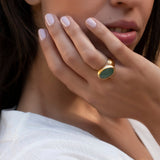 A gold ring with an oval, green stone. The ring is on a hand with light pink nail polish. The ring has a smooth, rounded band and a bezel setting for the stone. The background is blurred.