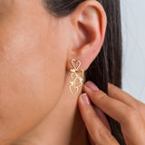Close-up of a woman wearing gold cut-out cascading heart drop earrings, with her hand gently touching the earring.