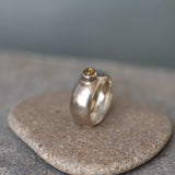 A handcrafted sterling silver ring with a wide, domed band and a polished finish. Set into the front of the band is a smooth, round light brown tourmaline gemstone in a bezel setting. The ring is photographed resting on a rough stone surface, highlighting its organic texture and contrast between the refined silver and earthy tones. The lighting emphasizes the gentle sheen of the silver and the warm depth of the gemstone.