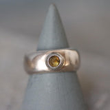 The image shows a handcrafted sterling silver ring with a wide, polished dome band. At its center is a smooth, round-cut light brown tourmaline gemstone, bezel-set for a clean, modern look. The silver surface is reflective and sleek, enhancing the warm, earthy tone of the tourmaline. The ring rests on a neutral background that highlights its shine and craftsmanship. The overall style is minimalist, elegant, and bold, ideal for women who love natural gemstone statement rings.