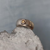 A handcrafted sterling silver ring with a wide, domed band and a polished finish. Set into the front of the band is a smooth, round light brown tourmaline gemstone in a bezel setting. The ring is photographed resting on a rough stone surface, highlighting its organic texture and contrast between the refined silver and earthy tones. The lighting emphasizes the gentle sheen of the silver and the warm depth of the gemstone.
