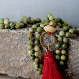 Close macro of the green jade beads on burgundy cord. The stones have subtle natural variations in tone and surface, with tight, secure knots visible between sections. The image captures the handcrafted quality and the matte sheen of the waxed cord.