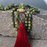 Detail shot of the tassel pendant on a jade mala necklace. Fine red-wine threads form a soft tassel attached to a burgundy waxed cord. Round green jade beads sit above the knot, revealing the handmade finish and smooth polish of each stone.