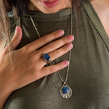 Close-up of a sterling silver jewelry set featuring a bold oval kyanite ring and a matching round kyanite pendant necklace. The inky blue gemstones show natural striations, bezel-set in polished silver. The necklace hangs on a fine silver chain beside the statement ring.