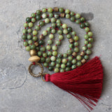 Flat lay of a boho mala necklace featuring polished green jade rounds on deep burgundy waxed cord with a coordinating red-wine tassel. The strand is long and lightweight, styled in a loose circle to highlight the color contrast between the cool green stones and warm cord.
