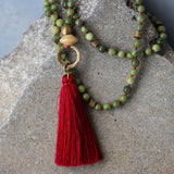 Lifestyle photo of the jade tassel necklace styled with a casual sweater. The cool green beads contrast with the warm burgundy cord and red-wine tassel. The long length and clasp-free construction make it suitable for everyday wear or meditation.
