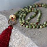 Close-up of a long beaded necklace made with smooth round green jade beads, strung on a burgundy red-wine waxed cord, ending in a matching red-wine thread tassel. The mala-style piece is clasp-free and designed to slip over the head, showing neat knots and a soft drape.