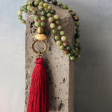 Overhead shot of the necklace coiled next to a small pouch. Round green jade beads, burgundy waxed cord, and a red-wine tassel are clearly visible. The image emphasizes portable, tangle-resistant design and the calming, natural look of the gemstone beads.