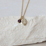 Close-up of a dainty gold leaf pendant necklace with a small deep-red garnet birthstone charm. The leaf has fine vein detailing and hangs from a slim gold chain, creating an elegant, minimalist look.