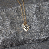 The necklace is arranged in a loose circle atop a matte gray stone surface. A small moonstone charm sits beside a satin-finished gold leaf pendant; the fine cable chain catches light subtly. The cool gray background makes the warm gold and opalescent gem stand out.