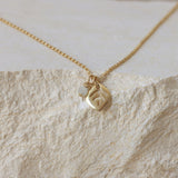 the gold-plated leaf pendant and moonstone charm placed on a light travertine/marble background. Soft, creamy tones complement the warm gold and the opalescent moonstone.