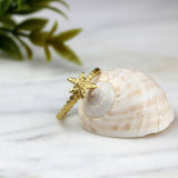 Delicate gold starfish ring leaning against four different seashells, styled in a coastal scene that enhances its ocean-inspired design