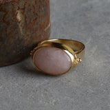 Gold rose quartz ring photographed on a grey stone background, showing the oval pink gemstone in a bezel setting.