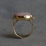 Gold rose quartz ring photographed on a grey stone background, showing the oval pink gemstone in a bezel setting.
