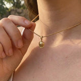 Dainty tiny floral gold pendant necklace with textured medallion charm and spiral bail on a gold chain, worn on the neck in warm natural sunlight.