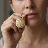 Close-up of a woman holding a round gold spiral pendant necklace near her lips, showing the engraved flowing swirl design on the coin-like charm.