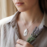 Woman wearing a handmade sterling silver rectangular pendant necklace with a raised lavender sprig, while holding dried lavender flowers against a soft neutral background.