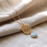 Close-up of a long gold necklace on soft linen fabric, featuring an openwork floral pendant with a round light-blue aquamarine stone in a gold bezel.