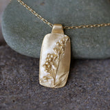 Close-up of a handmade gold rectangular pendant necklace featuring a raised lavender sprig relief, displayed against natural gray stones.