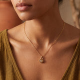 Woman wearing a tiny gold botanical pendant necklace with a round plant-inspired charm on a delicate chain, styled with a warm mustard top.