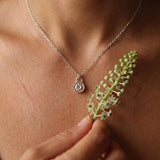 Woman wearing a tiny sterling silver botanical pendant necklace, holding a fresh green Phytolacca plant stem beside the charm.