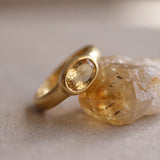 A gold ring with a large oval-shaped gemstone is resting on a piece of yellowish-brown rock.