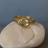 A gold ring with a large oval-shaped gemstone is displayed on a white rock.