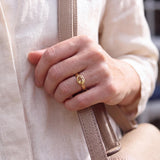 A close-up of a person's hand wearing a gold ring, with a beige purse strap visible.