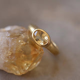 A gold ring with a large oval-shaped gemstone is displayed against a blurred background.