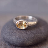 A silver ring with a large oval-shaped gemstone, possibly a citrine, is resting on a gray rock.
